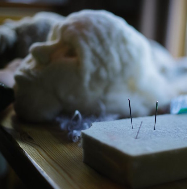Close up of needles stuck in a foam piece with needle felted face in the background.